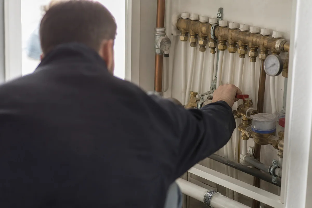 Technicien en combinaison qui coupe l’arrivée d’eau au compteur dans un placard technique, cuivre et PER apparents, lumière claire